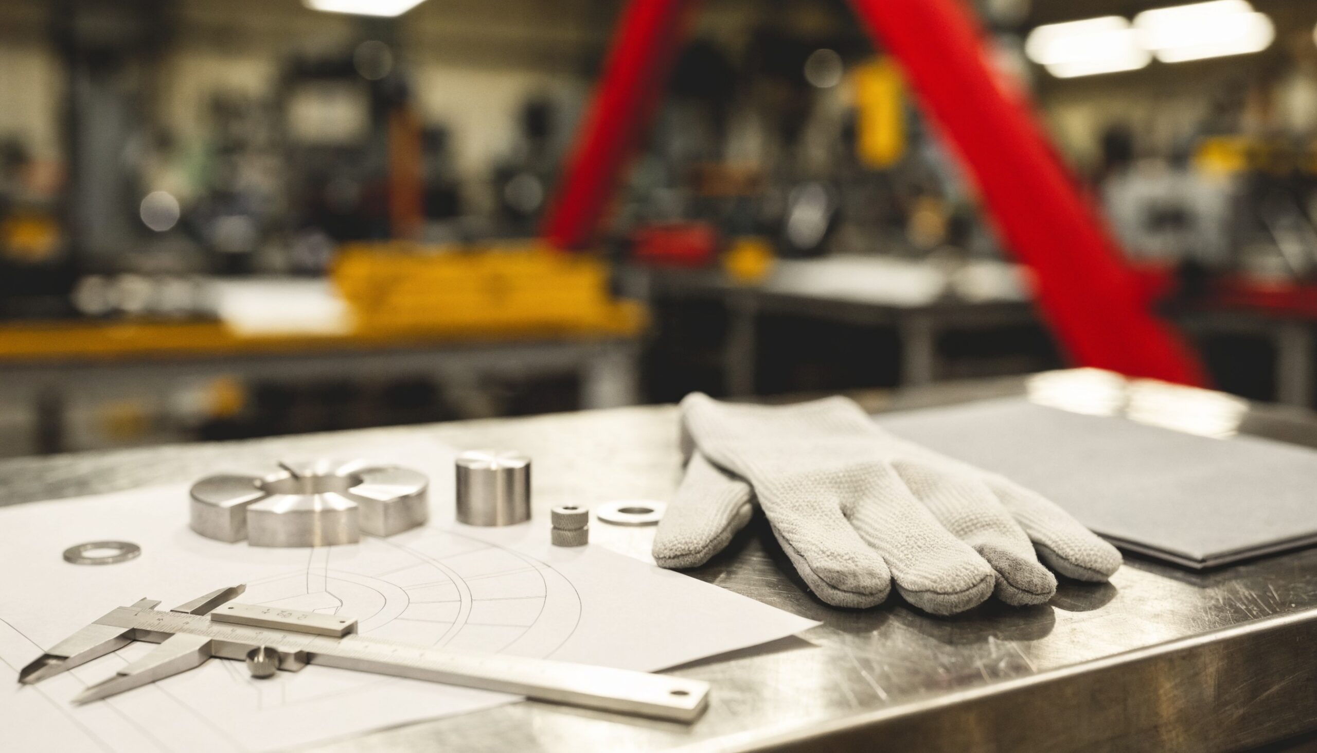 Werkstatt-Stillleben zur Bewerbung in der Metallbranche: Arbeitshandschuhe, Messschieber, technische Zeichnung und gefertigte Metallteile auf der Werkbank