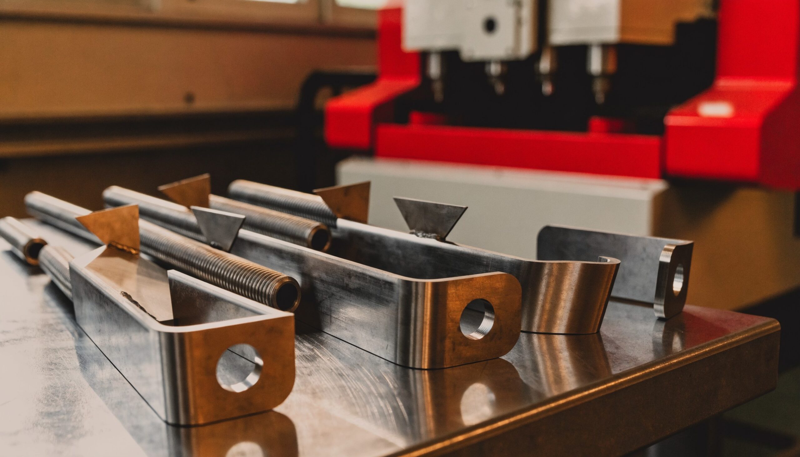 Gebogene Metallteile mit gequetschten Laschen, Bohrungen, Gewinde und angeschweißten Blechen vor einer Biegemaschine – Drahtbiegeteile mit Zusatzbearbeitung vom Hersteller